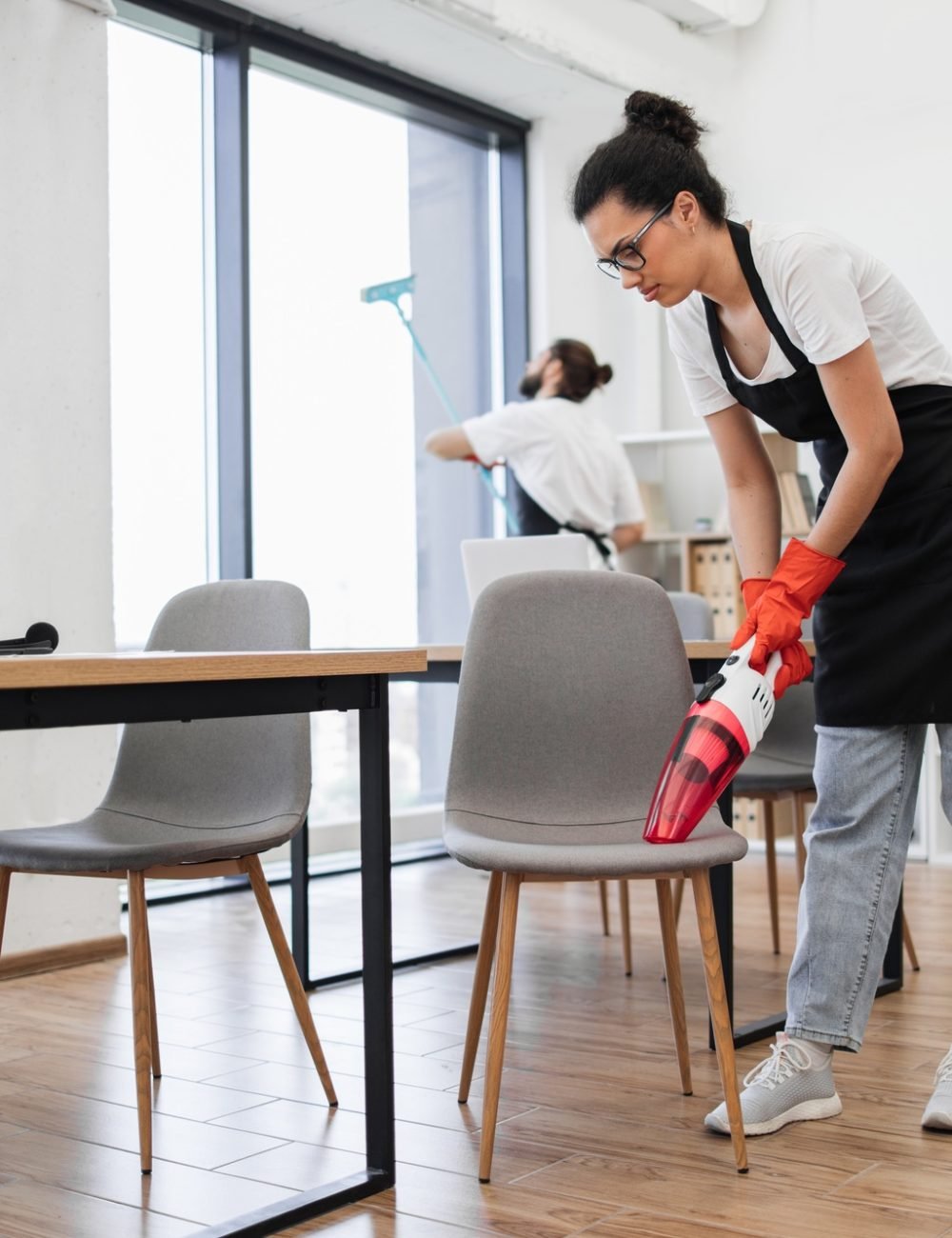 Multiethnic female cleaner vacuuming chair with portable vacuum cleaner.
